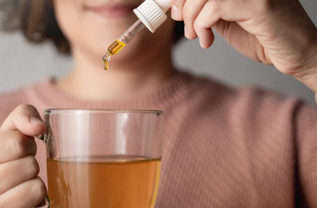 "A woman using an eye dropper to drop CBD oil into a glass of tea."